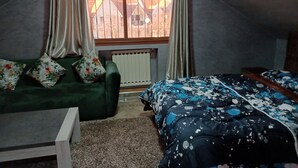 A bedroom with a sloped ceiling, a green sofa with floral cushions, a bed with a blue and white patterned duvet, and a window with metal bars and curtains. - Belle Vue Studios & Apartments (Ifrane)