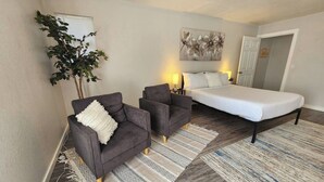 A bedroom with a bed, two armchairs, a potted plant, a painting, and a patterned rug. - Modern Retro Retreat (McKinleyville)