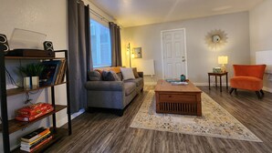 A living room with a grey sofa, a wooden coffee table, a small orange armchair, a bookshelf with books and a plant, a lamp, and a door with a mirror above it. - Modern Retro Retreat (McKinleyville)