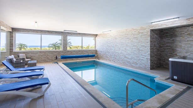 An indoor swimming pool with clear blue water, a hot tub, and a sun terrace with lounge chairs.