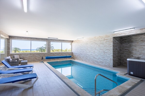 An indoor swimming pool with clear blue water, a hot tub, and a sun terrace with lounge chairs.