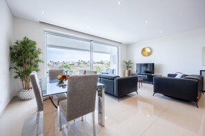 A modern living room with a glass dining table, two sofas, a TV, and a large window offering a view of buildings. - CoolHouses Algarve Lagos | Amor à Vida T3 (Lagos)