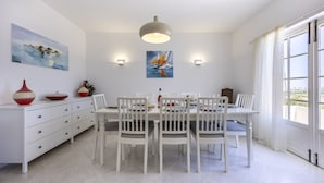 Zona pranzo con tavolo bianco apparecchiato per quattro, circondato da sedie bianche. Due lampade a parete e una plafoniera.