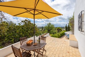 A patio with a table and chairs, a yellow umbrella, and a view of greenery and buildings.