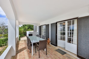 A covered patio with a glass table and wicker chairs, a view of greenery, and a sliding glass door. - CoolHouses Algarve Lagos | Casa Fernanda (Lagos)