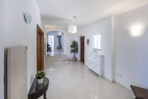 Un passadís amb una escala que condueix a una altra habitació, una petita taula consola blanca amb calaixos, una planta en test i una llum penjant.