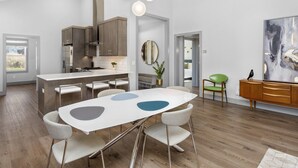 A modern kitchen with a central island, a dining area with a white table and grey chairs, and a living space with a green chair and a wooden cabinet. - Luxury Waterfront Getaway (Cullowhee)