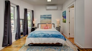 Un dormitorio con cama, dos mesitas de noche, una ventana con cortinas, un equipo de aire acondicionado y un cuadro en la pared.