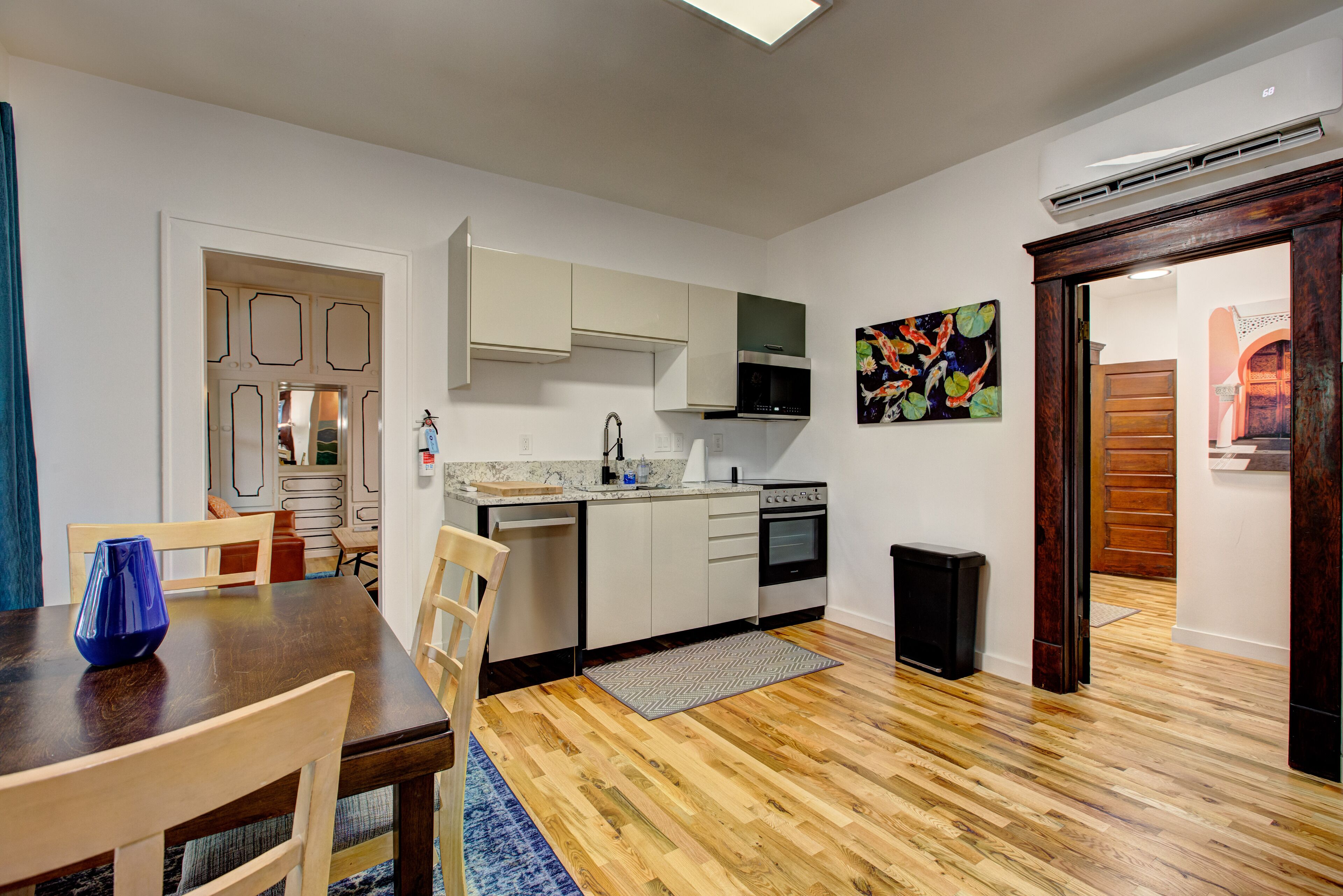 A compact kitchen with white cabinets, a microwave, and a stove. There is a dining table with chairs and a blue vase. The floor is wooden, and there is a rug in front of the table.