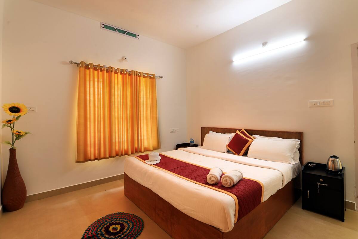 Una habitación de hotel ordenada con una cama grande, una mesita de noche, un jarrón con flores y una ventana con cortinas.