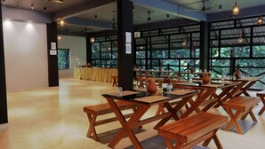 A spacious indoor area with wooden tables and benches, large windows offering a view of greenery outside.
