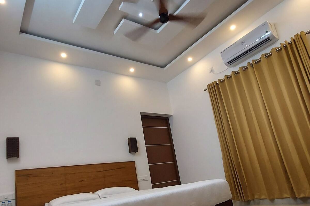 Un dormitorio con cama, ventilador de techo, iluminación empotrada, equipo de aire acondicionado y ventana con cortinas.