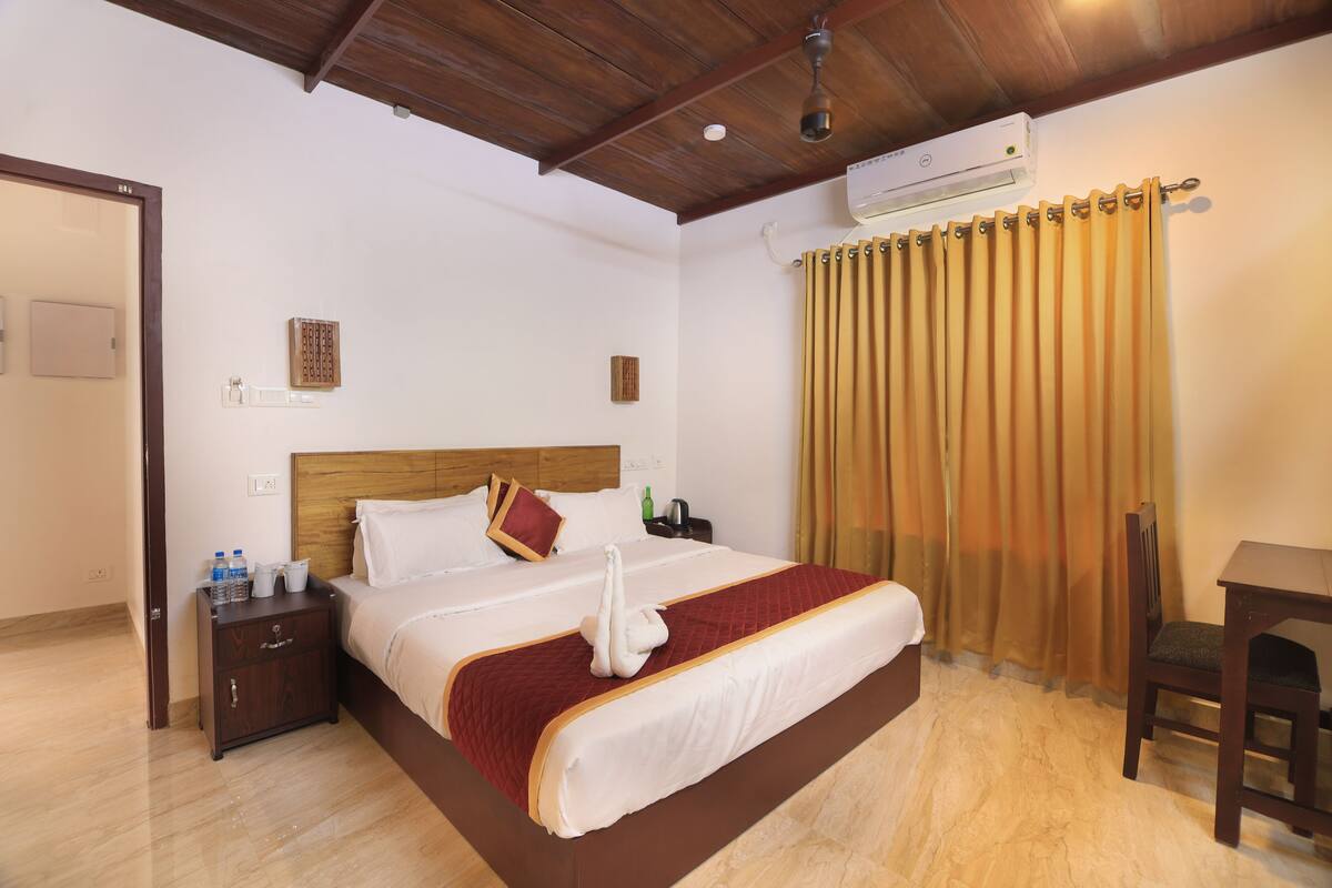 Una habitación de hotel con techo de madera, una cama con colcha roja y blanca, una mesita de noche de madera con botellas de agua, una silla de madera y una mesa.