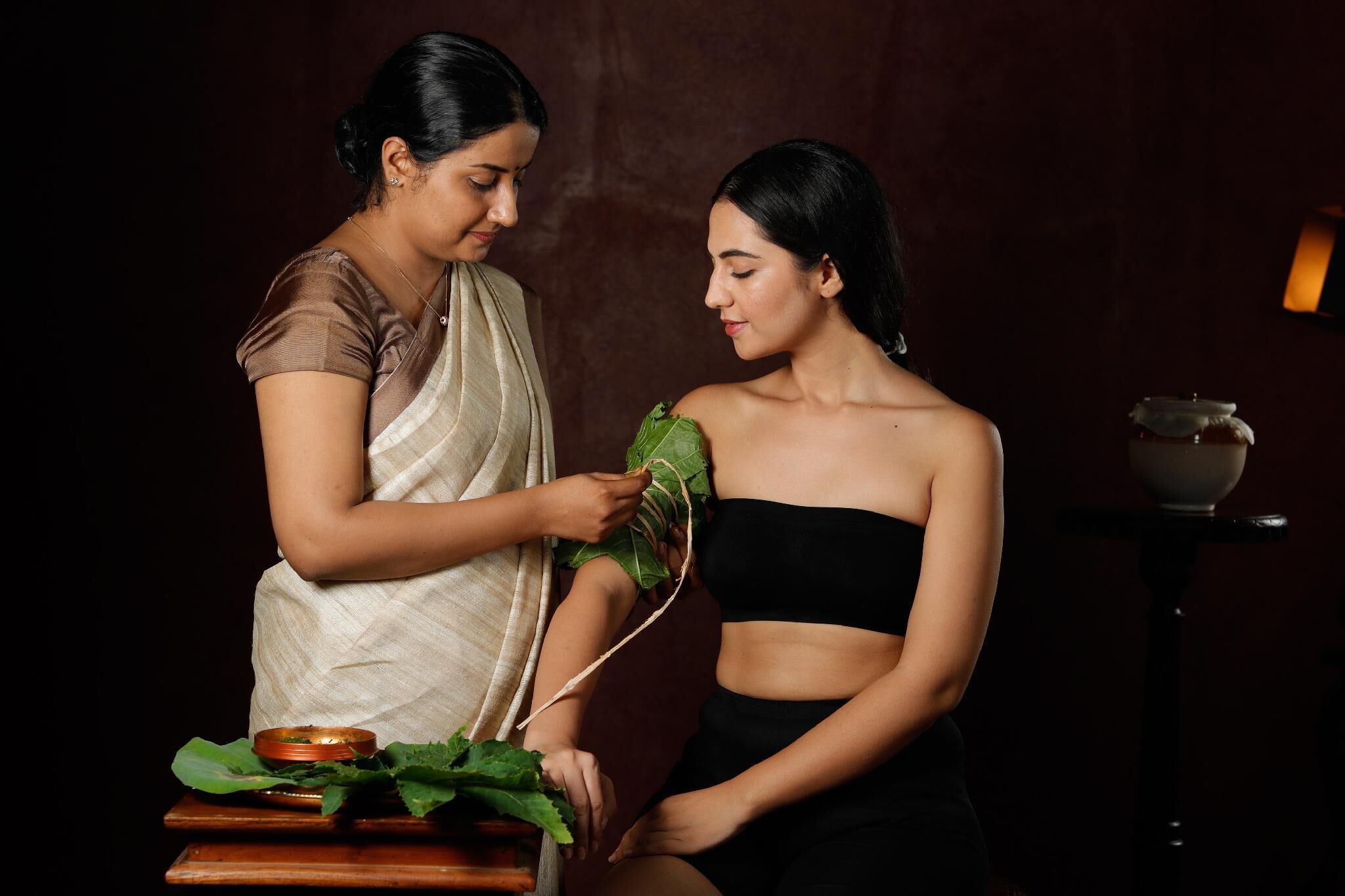 Two individuals in a room with a dark background, a small table with a bowl and leafy greens, and a lamp on a side table.