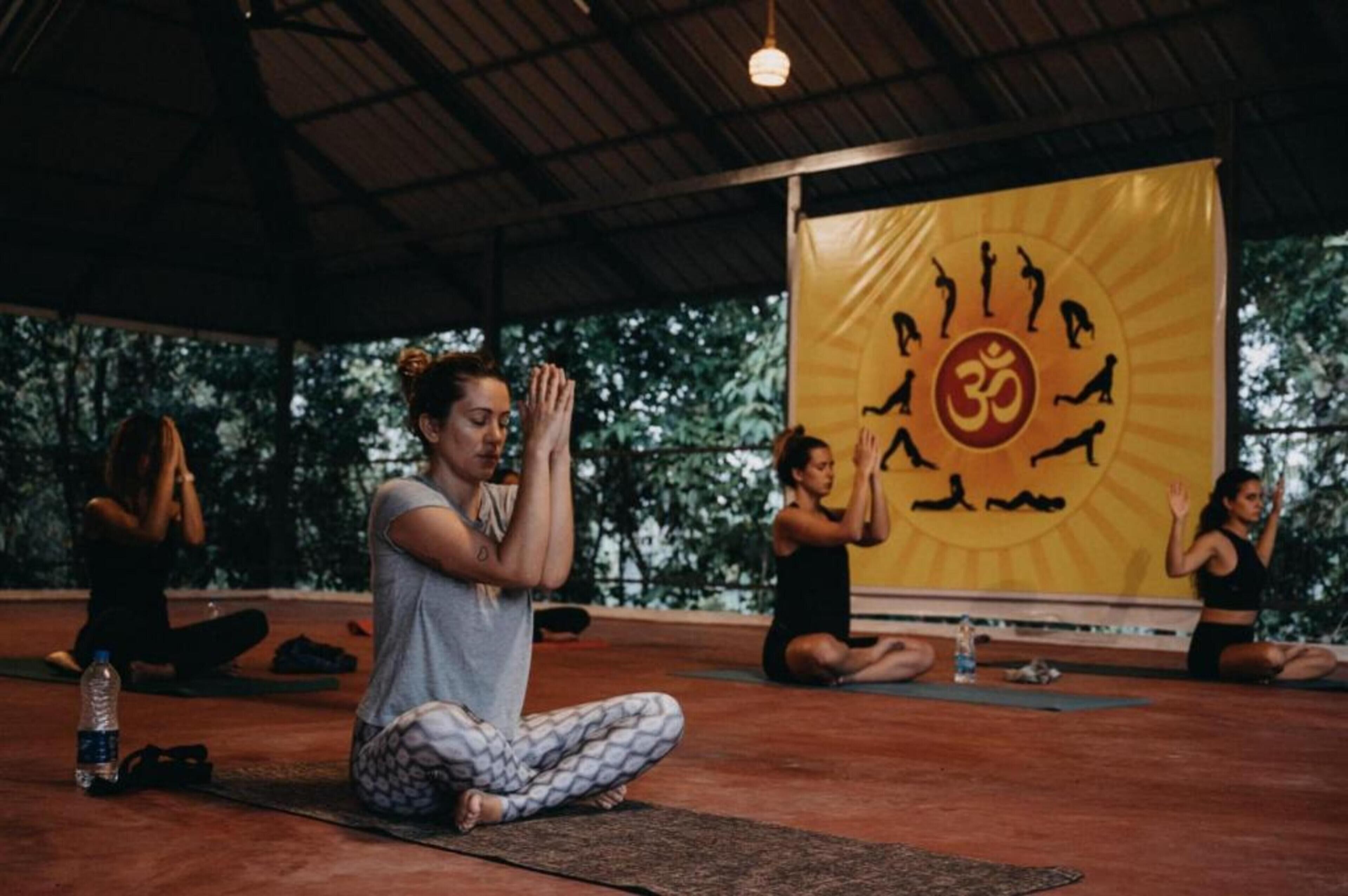 Una clase de yoga en una sala con un gran cartel amarillo que muestra un sol y posturas de yoga, techo alto y vistas a árboles fuera.