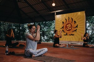 Una clase de yoga en una sala con un gran cartel amarillo que muestra un sol y posturas de yoga, techo alto y vistas a árboles fuera.