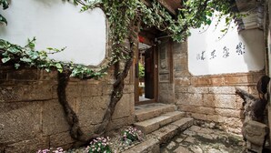 Ein traditioneller Innenhof mit Steinstufen, die zu einer Holztür führen, umrankt von Efeu und rosa Blüten.