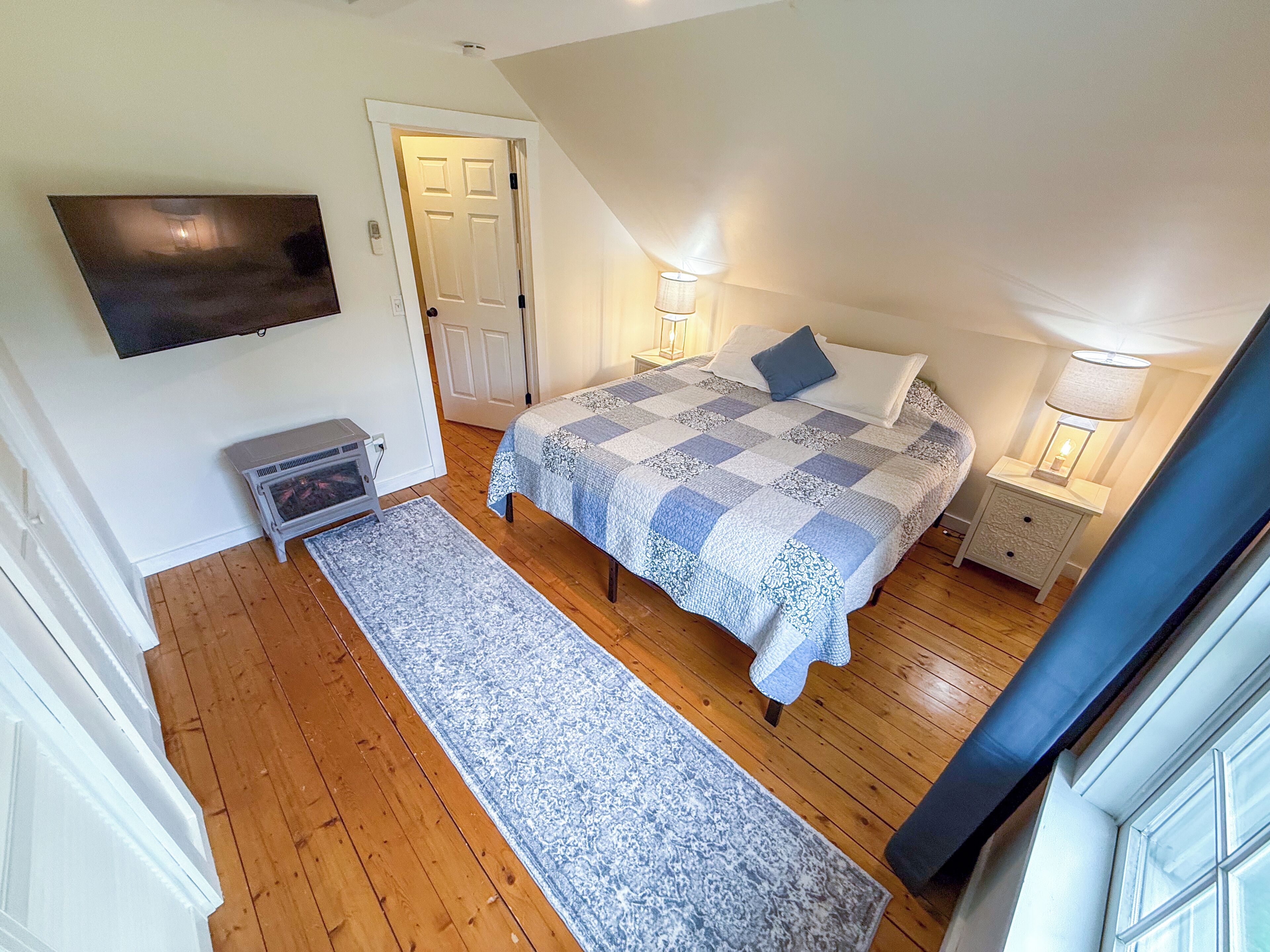 A bedroom with a bed, a nightstand, a TV mounted on the wall, a door, and a window.