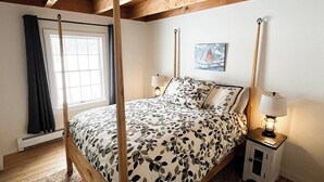 A four-poster bed with a floral bedspread, a bedside table with a lamp, and a window with curtains.
