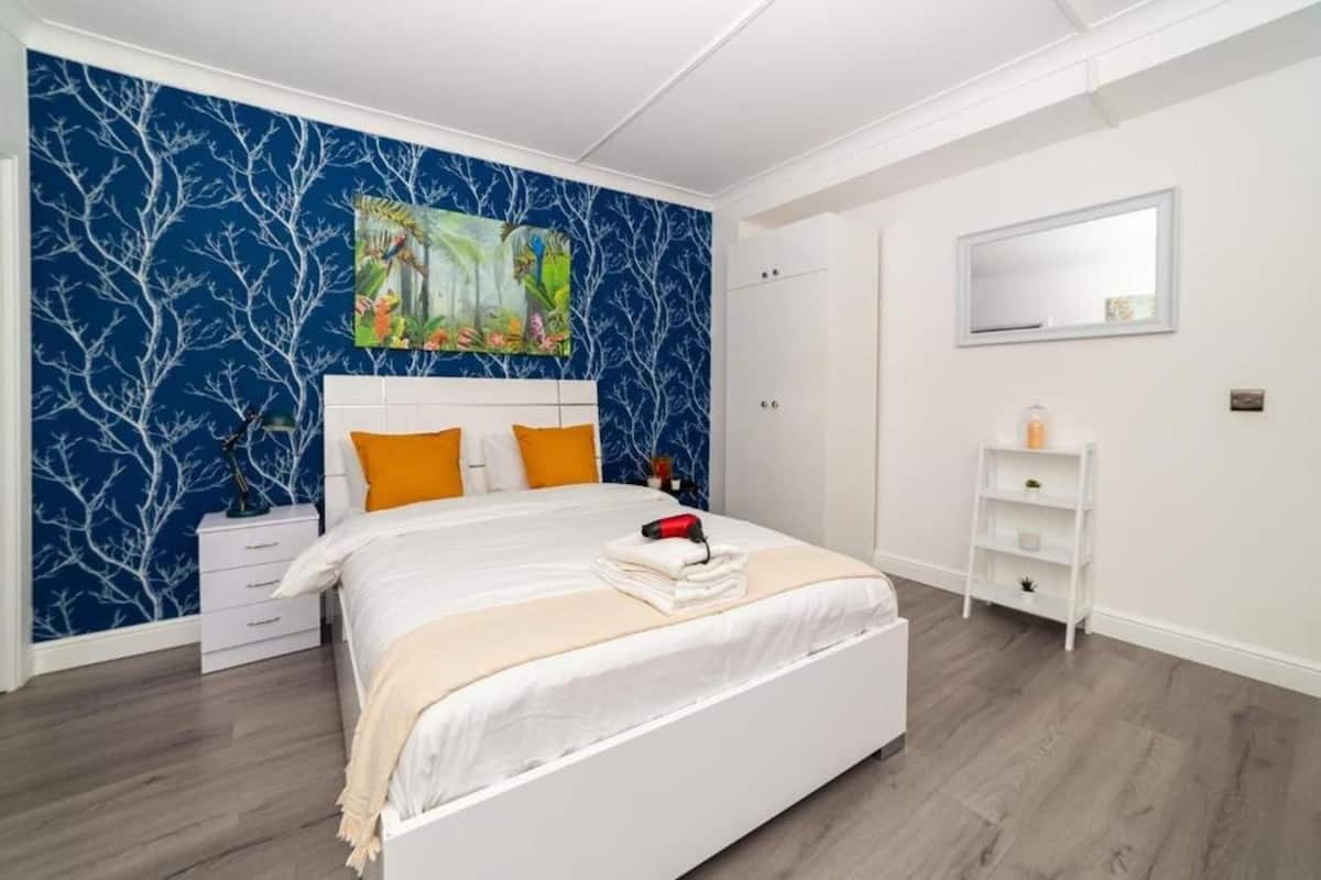 Een slaapkamer met een bed, witte nachtkastjes, een lampje op een nachtkastje, een witte dressoir, een spiegel en een muur met een blauw boompatroon.