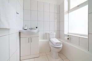 A modern bathroom with white tiles, a toilet, a sink, and a window. - Diana Hotel Near Wembley Self Serviced Apartments (London)