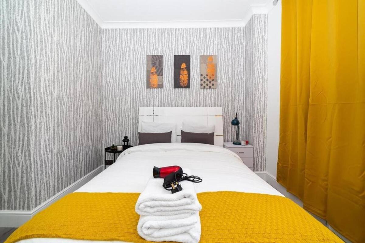 Een keurig opgemaakt bed met een gele deken en witte handdoeken, een rode haardroger en een nachtkastje met een lamp.