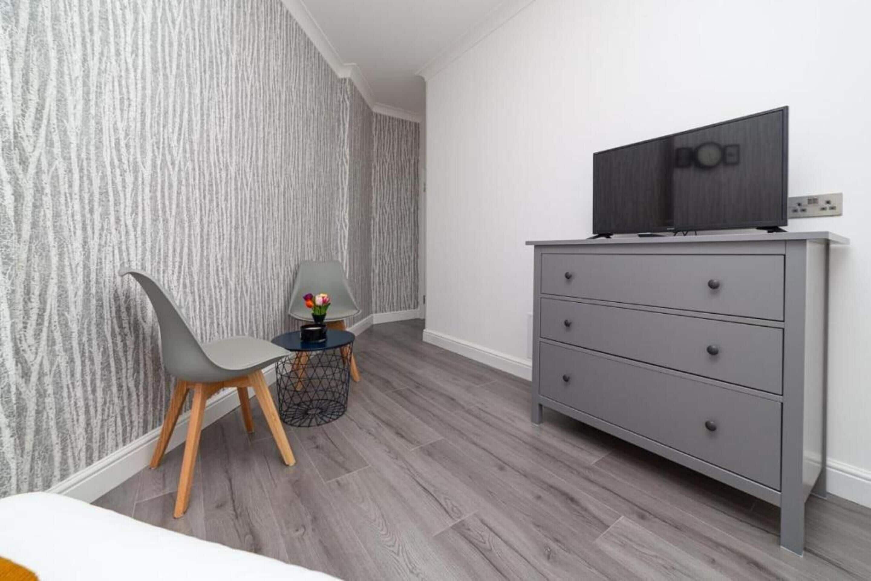 Ein modernes Schlafzimmer mit einer grauen Kommode, einem Fernseher darauf, einem Sessel und einem kleinen runden Tisch mit Blumen in einer Vase.