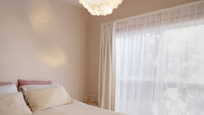 Quarto com cama, mesa de cabeceira e janela com cortinas leves.