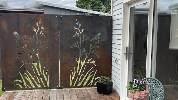 Um deck de madeira com grade de metal decorada com motivos de plantas, uma porta de vidro e uma mesa com um vaso.