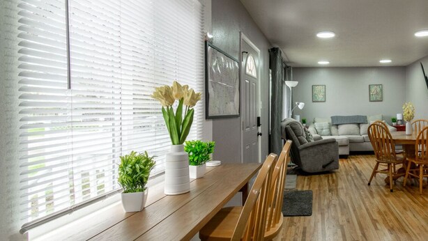 A modern living room with a wooden dining table, chairs, a grey sofa, and a coffee table. There are potted plants on the windowsill and a framed picture on the wall.