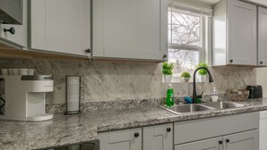 A modern kitchen with white cabinets, a granite countertop, a coffee maker, a sink, and a window with a view of trees.
