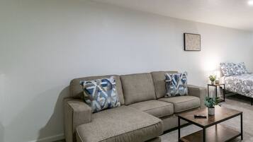 Una sala de estar con un sofá seccional color beige, dos almohadones estampados, una pequeña mesa de centro de madera y un cuadro en la pared.