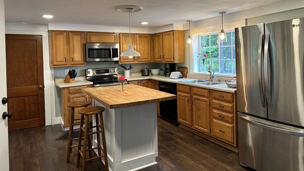 A modern kitchen with wooden cabinets, stainless steel appliances, and a central island with stools. - Downtown Meredith Village Cape (Meredith)