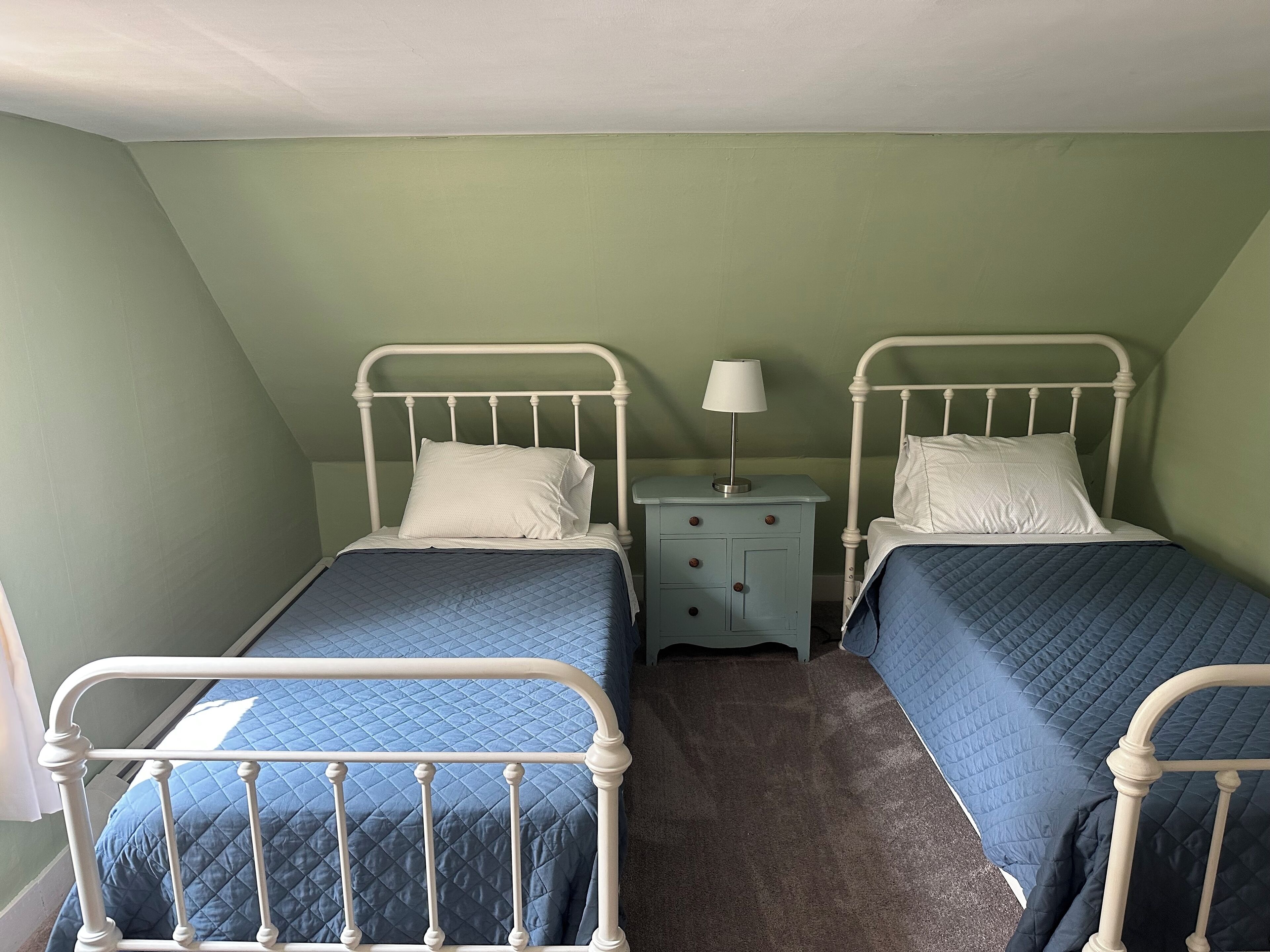 A twin room with two beds, each with a white headboard and blue bedding. There is a small bedside table with a lamp between the beds.