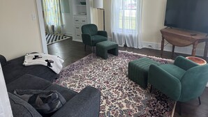 A living room with a sofa, two green armchairs, a patterned rug, a television on a wooden stand, and a window with sheer curtains. - Downtown Meredith Village Cape (Meredith)