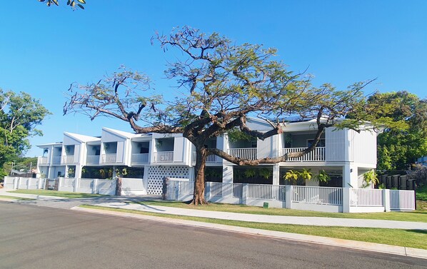 A row of two-story townhouses with balconies, a large tree in front, and a clear blue sky.