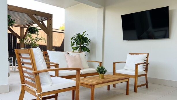 A modern outdoor seating area with wooden chairs and a table, surrounded by potted plants and a TV mounted on the wall. - One on Harris Apartments (Wellington Point)
