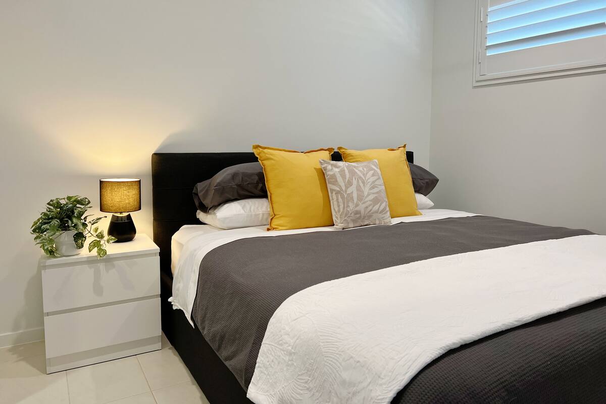 A neatly made bed with a black headboard, white and gray bedding, and multiple pillows in yellow and white. A bedside table with a lamp and a potted plant is visible.