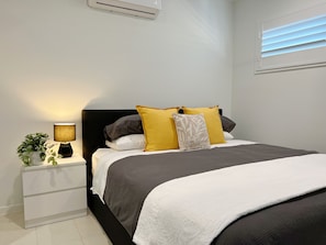 A neatly made bed with a black headboard, white and gray bedding, and multiple pillows in yellow and white. A bedside table with a lamp and a potted plant is visible.