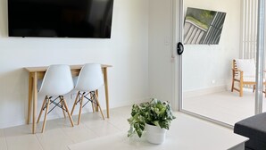A modern living room with a flat-screen TV, a wooden desk with two white chairs, a potted plant, and a sliding glass door leading to an outdoor area with a wooden chair and a cushioned seat. - One on Harris Apartments (Wellington Point)