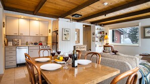 A cozy kitchen with wooden beams, a dining table set for two, and a living area with a sofa and a window.