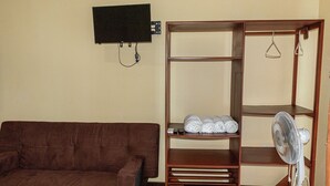 A room with a brown sofa, a wooden wardrobe with shelves, a white fan, and a mounted television. - San Alejo Zero (Charapoto)