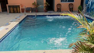 Una piscina rectangular con agua cristalina azul, rodeada por un solárium con baldosas y plantas en macetas.