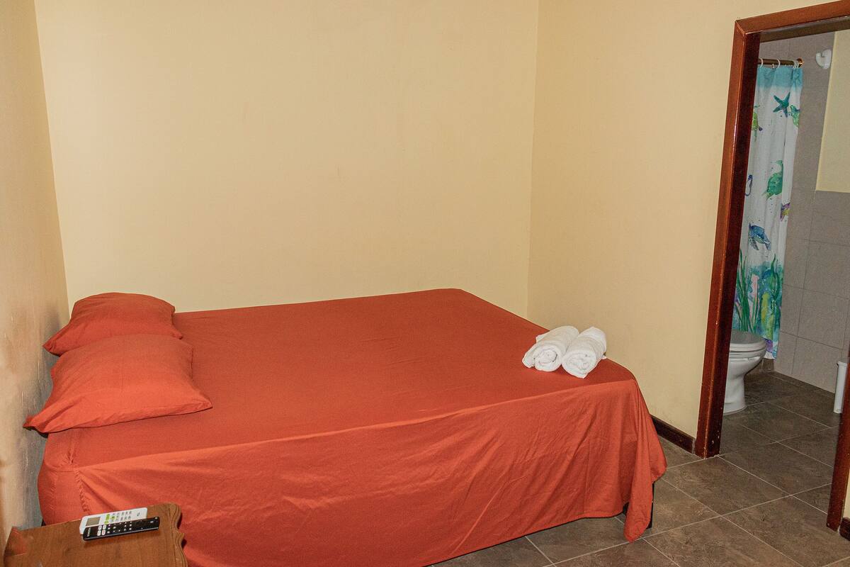 A small, single-bed room with a red bedspread, a remote control on a wooden side table, and a set of towels on the bed.