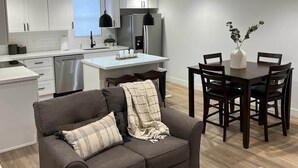 Een moderne keuken met witte kastjes, een eethoek met een houten tafel en stoelen, en een grijze bank met een plaid.