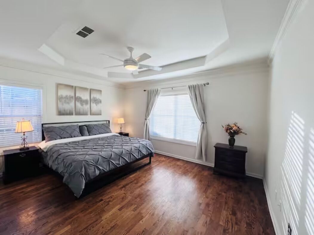 Une chambre à coucher comprenant un lit, des tables de chevet, un ventilateur de plafond et une fenêtre avec des rideaux.