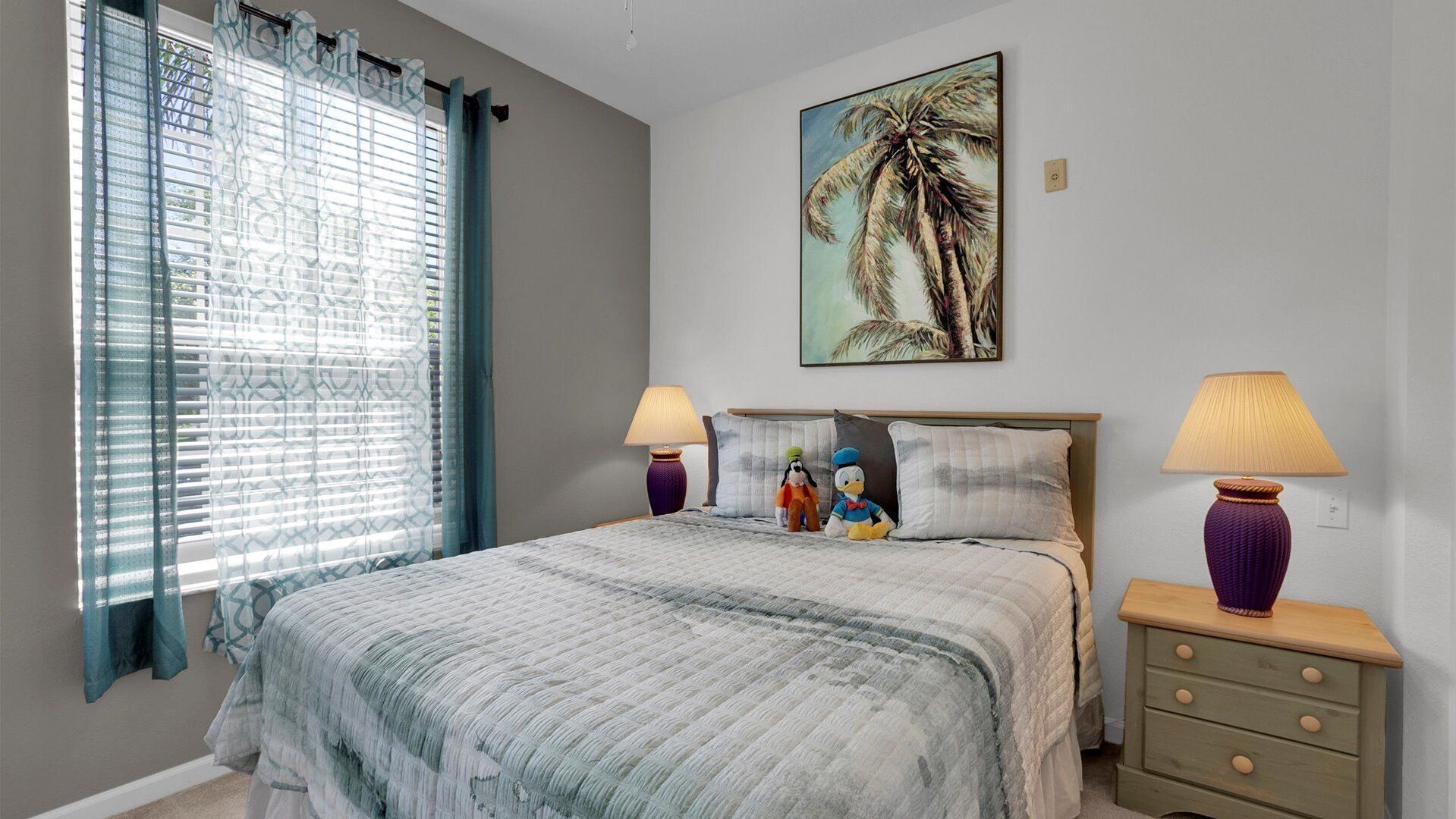 A bedroom with a bed, two nightstands, a lamp on each, a window with curtains, and a painting on the wall.