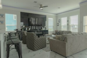 A modern living room with a large flat-screen TV, comfortable seating, and a ceiling fan. - Brand New Custom Construction! (Freeport)