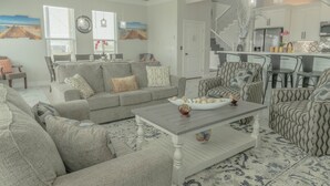Un vaste salon avec un canapé gris, des fauteuils à motifs et une table basse. Un escalier mène à une cuisine équipée de meubles blancs et à un coin repas.