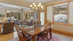Un vaste salon doté d’une grande table à manger, de chaises en bois et d’un lustre. De grandes fenêtres équipées de stores, un ventilateur de plafond et une vue sur un paysage enneigé complètent l’ensemble.